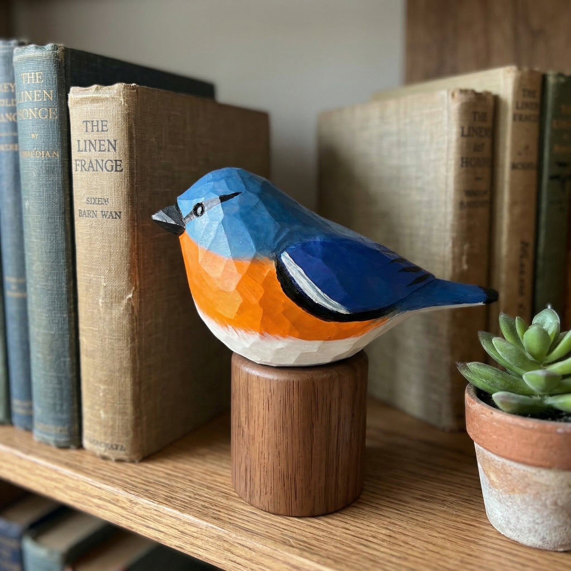 A geometric, faceted wooden Eastern Bluebird sculpture in blue, orange, and white, mounted on a cylindrical wooden base and displayed on a wooden shelf next to vintage books and a small potted succulent.