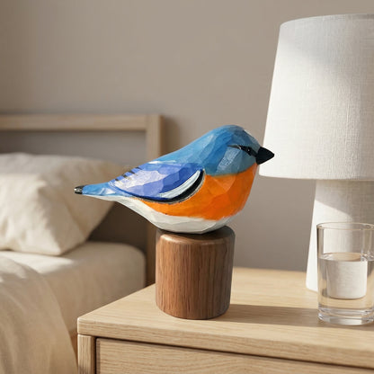 A geometric, faceted bird sculpture in blue, orange, and white sits on a wooden cylindrical base on a light wood bedside table, with a lamp and glass of water in a softly lit bedroom setting.