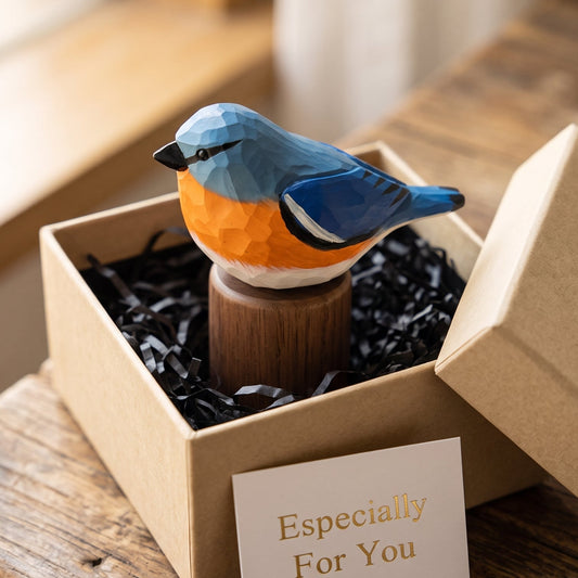 A small geometric Eastern Bluebird figurine in blue, orange, and white sits on a wooden stand inside an open kraft gift box with black shredded paper. A card reading 'Especially For You' is placed in front, and the box lid rests beside it on a rustic wooden table.
