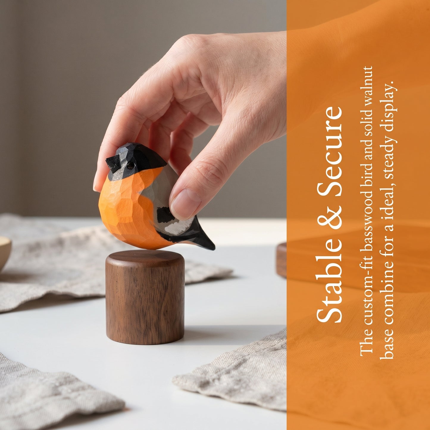 A hand holds a small carved Eurasian bullfinch-shaped object above a round wooden base on a tabletop. The product appears to be a decorative bird display or stand, with an orange, black, and gray carved bird and a medium brown wooden base.