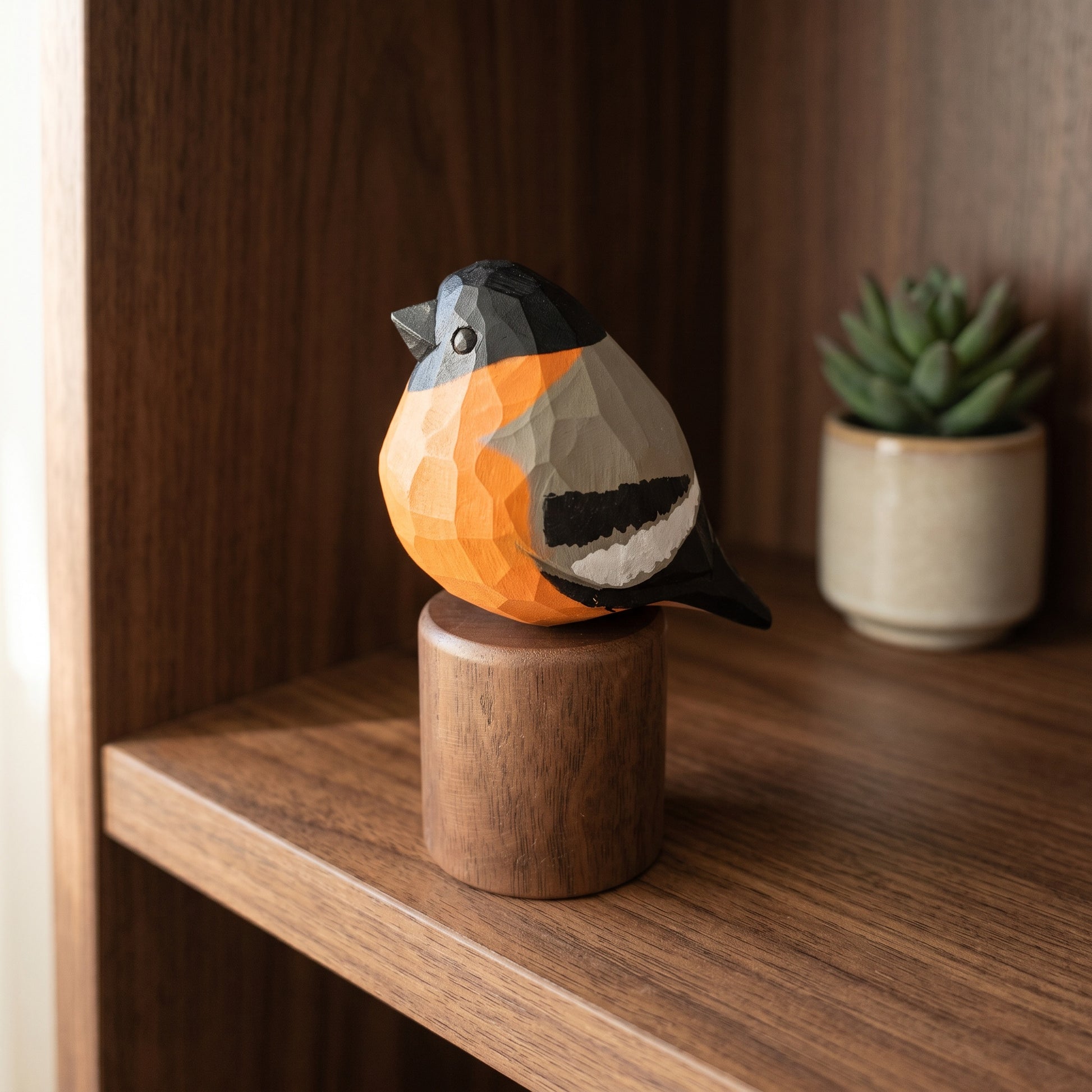 A geometric Eurasian bullfinch figurine with a faceted orange, black, gray, and white design, perched on a cylindrical wooden base. The figurine is displayed on a wooden shelf next to a small potted succulent.