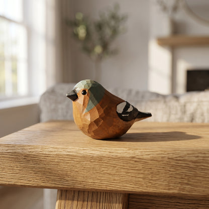 A low-poly wooden Eurasian chaffinch figurine with a geometric faceted design, resting on a light oak table in a softly blurred living room setting.