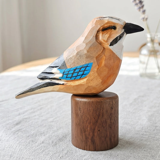 A hand-carved wooden Eurasian jay figurine with geometric facets, featuring a warm orange-brown body, white head, black beak, gray wings, and a bright blue wing accent, mounted on a cylindrical wooden base. The bird is displayed on a light gray linen-covered table with a blurred glass vase of lavender in the background.
