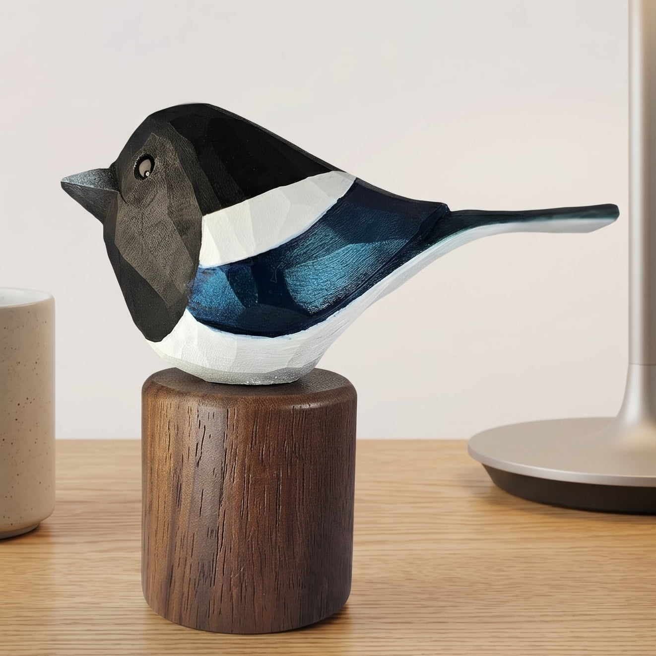 A handcrafted wooden Eurasian magpie figurine with painted black, white, and blue details, mounted on a dark wood cylindrical base and displayed on a light wood tabletop next to a ceramic mug and a table lamp.