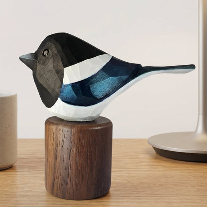 A handcrafted wooden Eurasian magpie figurine with painted black, white, and blue details, mounted on a dark wood cylindrical base and displayed on a light wood tabletop next to a ceramic mug and a table lamp.