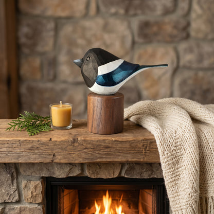 A painted Eurasian magpie figurine perched on a wooden stump base, displayed on a rustic wooden mantel in front of a stone fireplace with a candle, evergreen sprig, and cream knit throw blanket nearby.