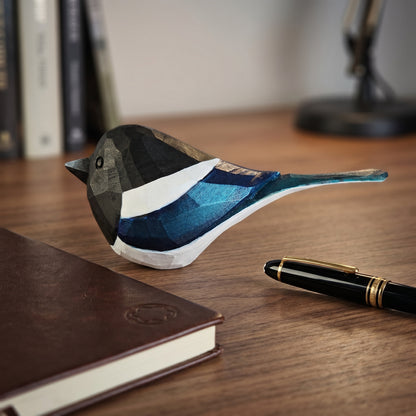 A geometric Eurasian magpie figurine with faceted black, white, and blue facets, styled on a wooden desk next to a brown notebook and a black pen.