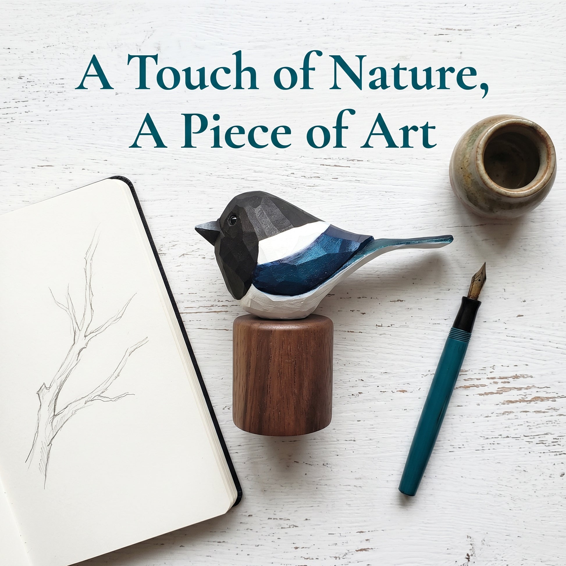 A geometric Eurasian magpie figurine with a black, white, and blue patterned body, mounted on a round wooden base. The figurine is styled in a flat-lay arrangement on a white wood background alongside a notebook with a branch sketch, a teal fountain pen, and a small ceramic pot.