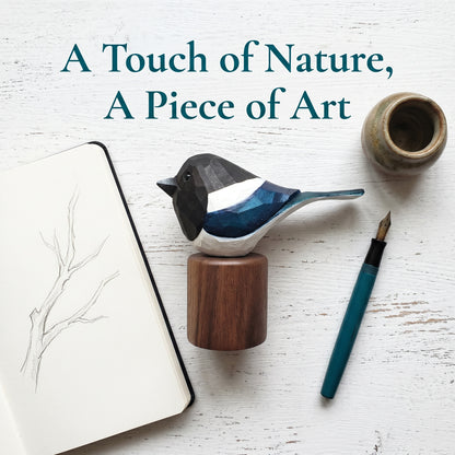 A geometric Eurasian magpie figurine with a black, white, and blue patterned body, mounted on a round wooden base. The figurine is styled in a flat-lay arrangement on a white wood background alongside a notebook with a branch sketch, a teal fountain pen, and a small ceramic pot.