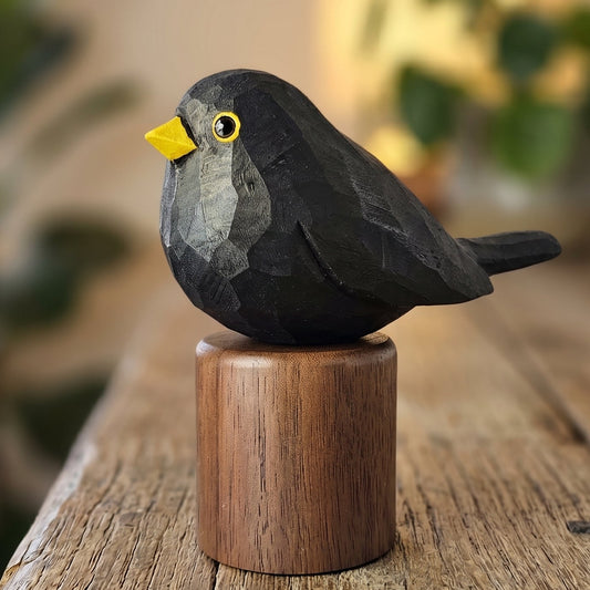 A small carved Eurasian Blackbird figurine with a black body, yellow beak, and yellow eye, perched on a round wooden base on a rustic wooden table with a softly blurred indoor background.
