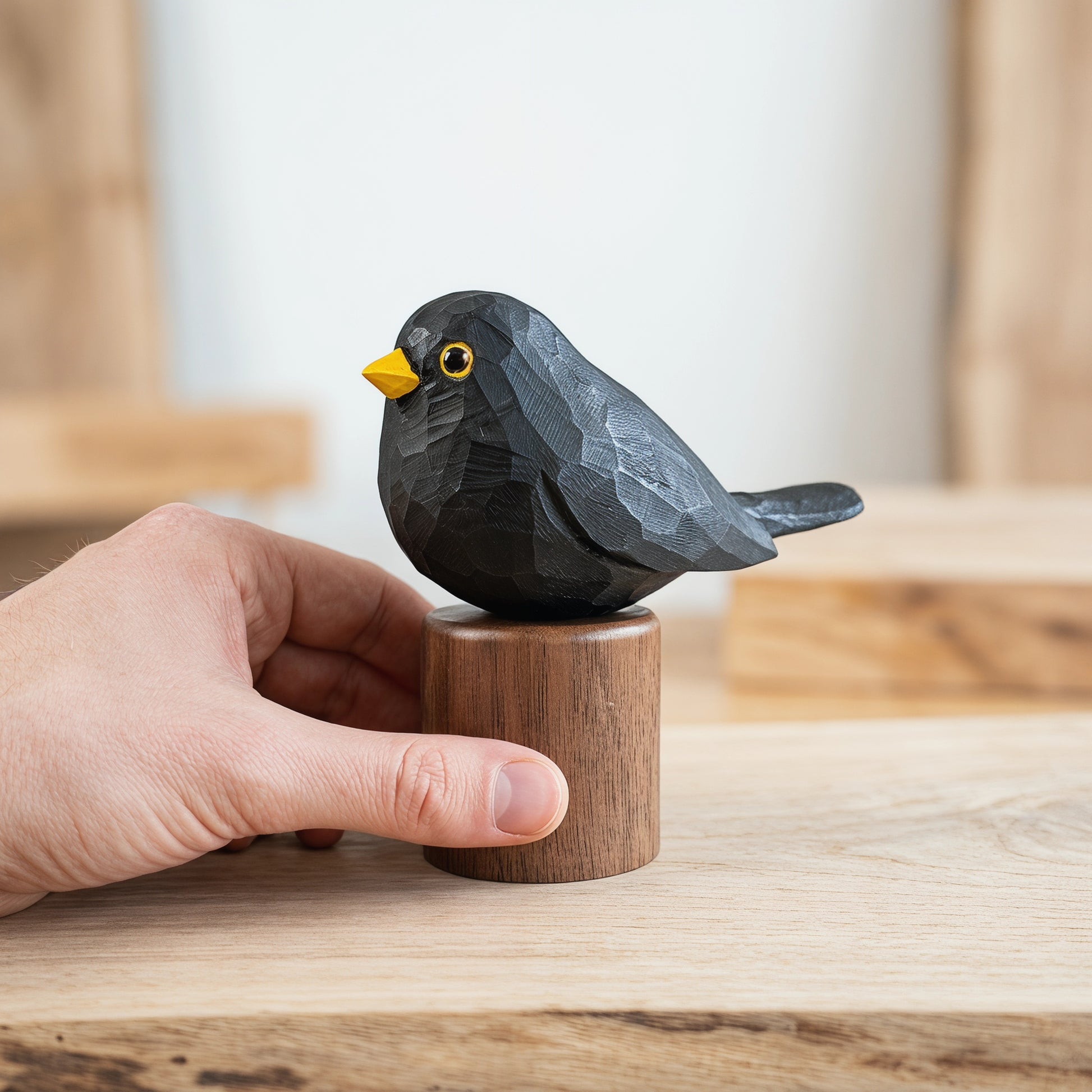 A small carved Eurasian Blackbird figurine in dark charcoal gray with a bright yellow beak and yellow eye, mounted on a round wooden base, being held by a person's hand on a light wood surface.
