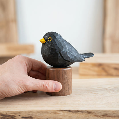 A small carved Eurasian Blackbird figurine in dark charcoal gray with a bright yellow beak and yellow eye, mounted on a round wooden base, being held by a person's hand on a light wood surface.