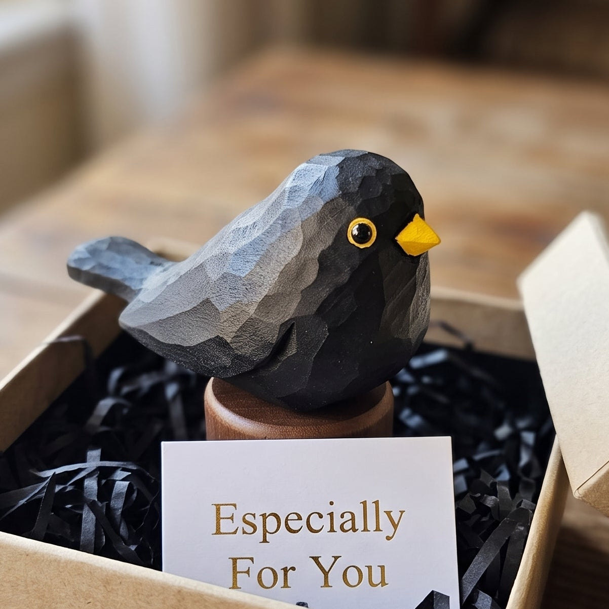 A small Eurasian Blackbird figurine in a brown pot sits inside a kraft gift box with black shredded paper filler and a card that reads 'Especially For You,' placed on a wooden table.