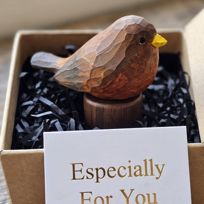 A carved wooden Eurasian Blackbird figurine with a yellow beak displayed in a kraft gift box filled with black shredded paper, with a white card reading 'Especially For You' in front.