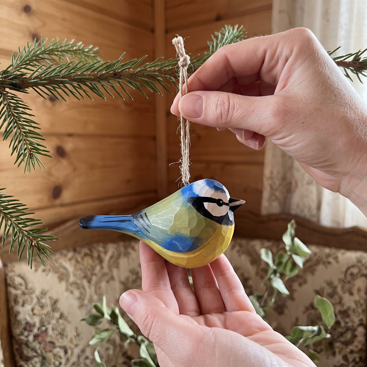 A handcrafted wooden Eurasian blue tit ornament painted like a blue tit, hanging from a pine branch held by a person's hand in a cozy indoor setting.