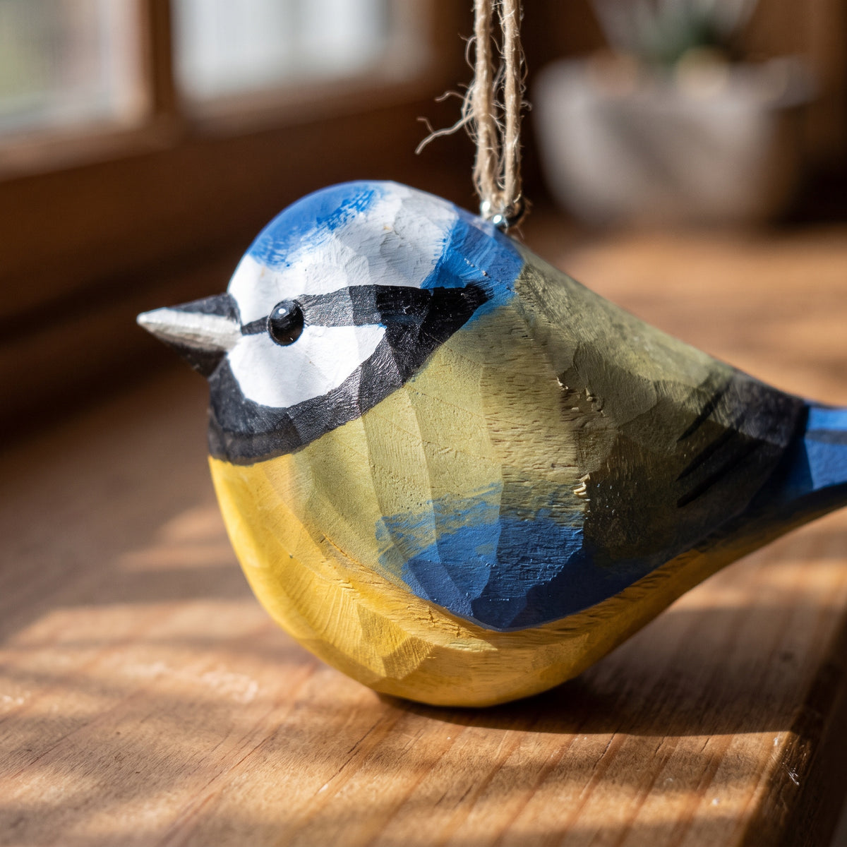 A handcrafted wooden Eurasian blue tit ornament painted in blue, yellow, black, and white, hanging by a twine loop on a wooden surface.