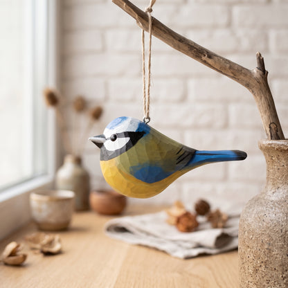 A handcrafted wooden Eurasian blue tit ornament in blue, yellow, and green hangs from a rustic branch on a wooden table, with a blurred indoor background featuring a vase, small bowls, and a white brick wall.
