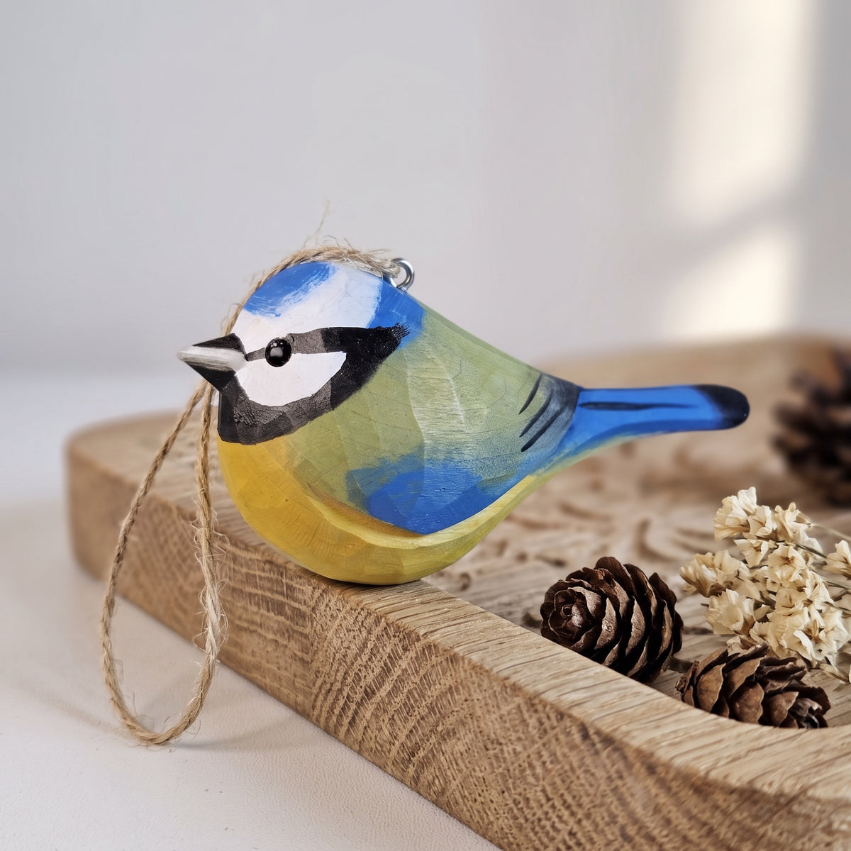 A hand-painted wooden Eurasian blue tit ornament resembling a blue tit, featuring blue, yellow, green, black, and white colors, with a jute hanging loop. It is displayed on a rustic wooden tray with pinecones and dried flowers.