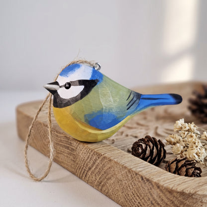 A hand-painted wooden Eurasian blue tit ornament resembling a blue tit, featuring blue, yellow, green, black, and white colors, with a jute hanging loop. It is displayed on a rustic wooden tray with pinecones and dried flowers.