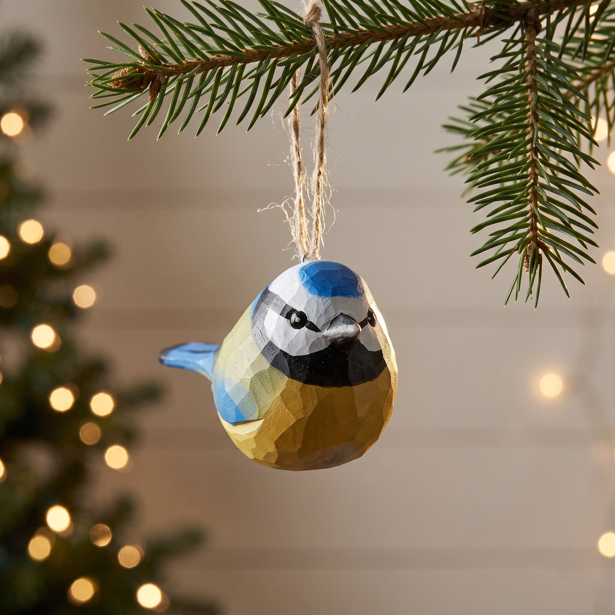 A faceted Eurasian blue tit-shaped Christmas ornament in blue, yellow, black, and white hangs from a pine branch with twine, with blurred holiday lights in the background.