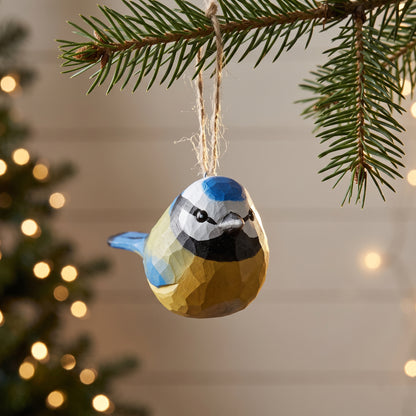 A faceted Eurasian blue tit-shaped Christmas ornament in blue, yellow, black, and white hangs from a pine branch with twine, with blurred holiday lights in the background.