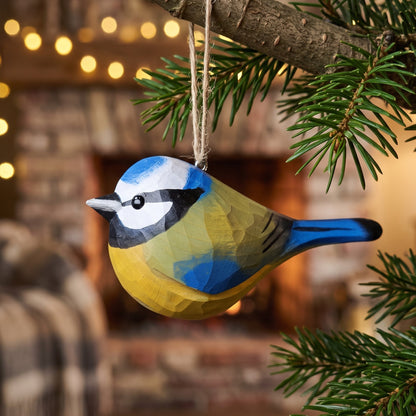 A handcrafted wooden Eurasian blue tit Christmas ornament, painted in the colors of a blue tit with yellow, blue, black, and white, hanging from a tree branch in front of a cozy, blurred fireplace background.