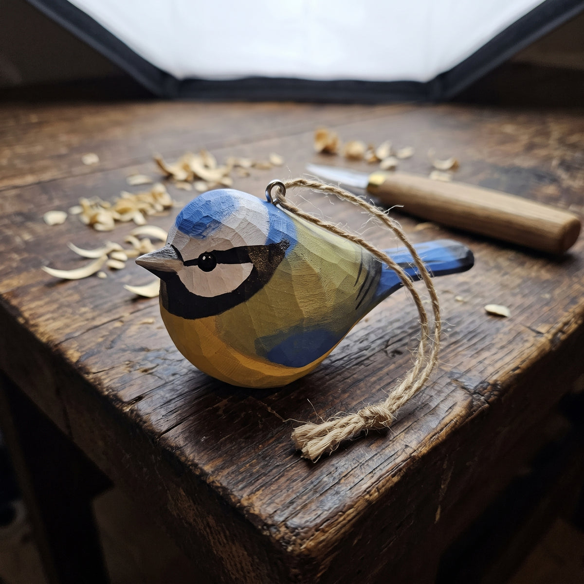 A handcrafted wooden Eurasian blue tit ornament painted in blue, yellow, and black, resting on a rustic wooden workbench with carving tools and wood shavings nearby.