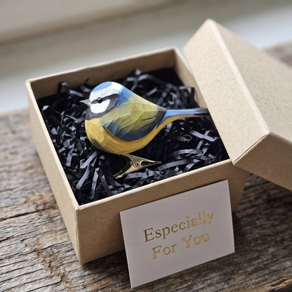 A hand-carved wooden Eurasian Blue Tit clip-on ornament resting in an open kraft gift box filled with black shredded paper, featuring a light blue cap, yellow underbelly, black eye-stripe, and a small card reading "Especially For You" placed in the foreground.