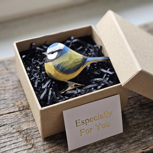 A hand-carved wooden Eurasian Blue Tit clip-on ornament resting in an open kraft gift box filled with black shredded paper, featuring a light blue cap, yellow underbelly, black eye-stripe, and a small card reading "Especially For You" placed in the foreground.