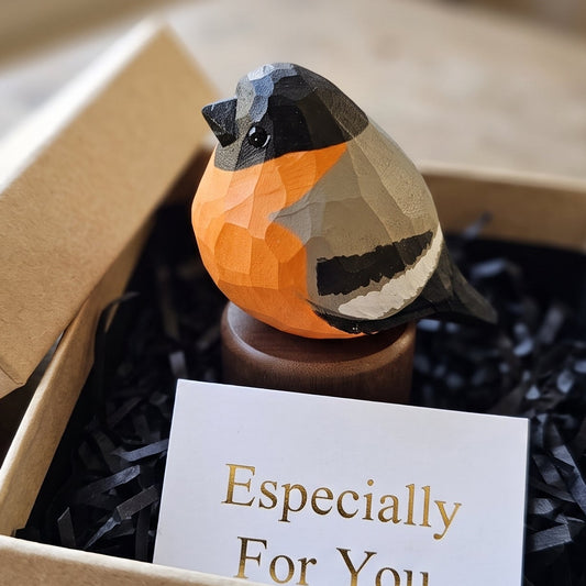 A geometric Eurasian bullfinch figurine with orange, black, gray, and white facets sits in a kraft gift box filled with black shredded paper and a card that reads 'Especially For You'.