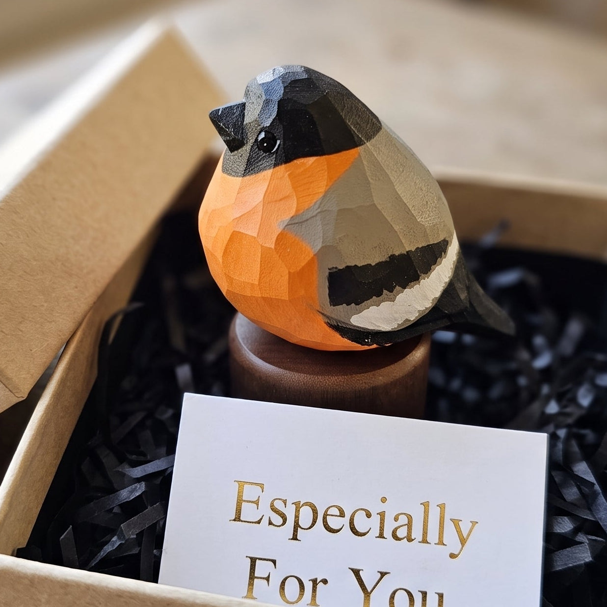 A geometric Eurasian bullfinch figurine with orange, black, gray, and white facets sits in a kraft gift box filled with black shredded paper and a card that reads 'Especially For You'.