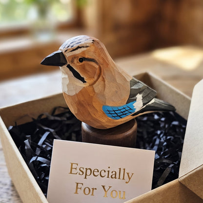 A handcrafted wooden Eurasian jay figurine with blue and black painted accents sits in a brown kraft gift box filled with black shredded paper and a small card reading 'Especially For You.' The box is placed on a rustic wooden table with a softly blurred indoor background.