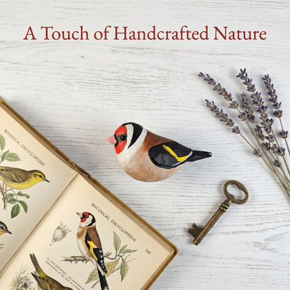 A handcrafted wooden European Goldfinch figurine with a red, black, white, and yellow painted pattern, styled on a white wood background with an open botanical dictionary, a vintage key, and dried lavender. The text 'A Touch of Handcrafted Nature' is written at the top.