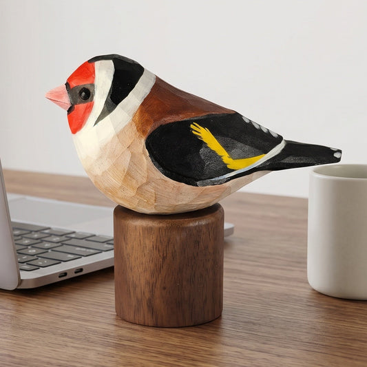 A hand-painted wooden European Goldfinch figurine perched on a cylindrical wooden base, styled on a wooden desk next to a laptop, mug, and coffee cup.