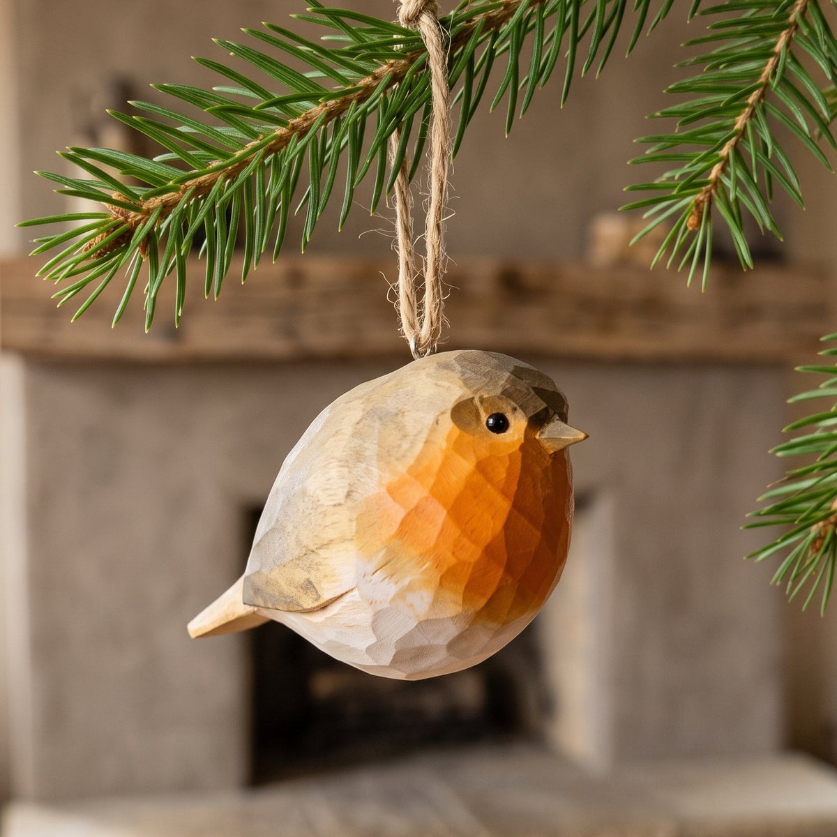 A faceted wooden robin bird ornament hangs from a pine branch in front of a softly blurred fireplace background.