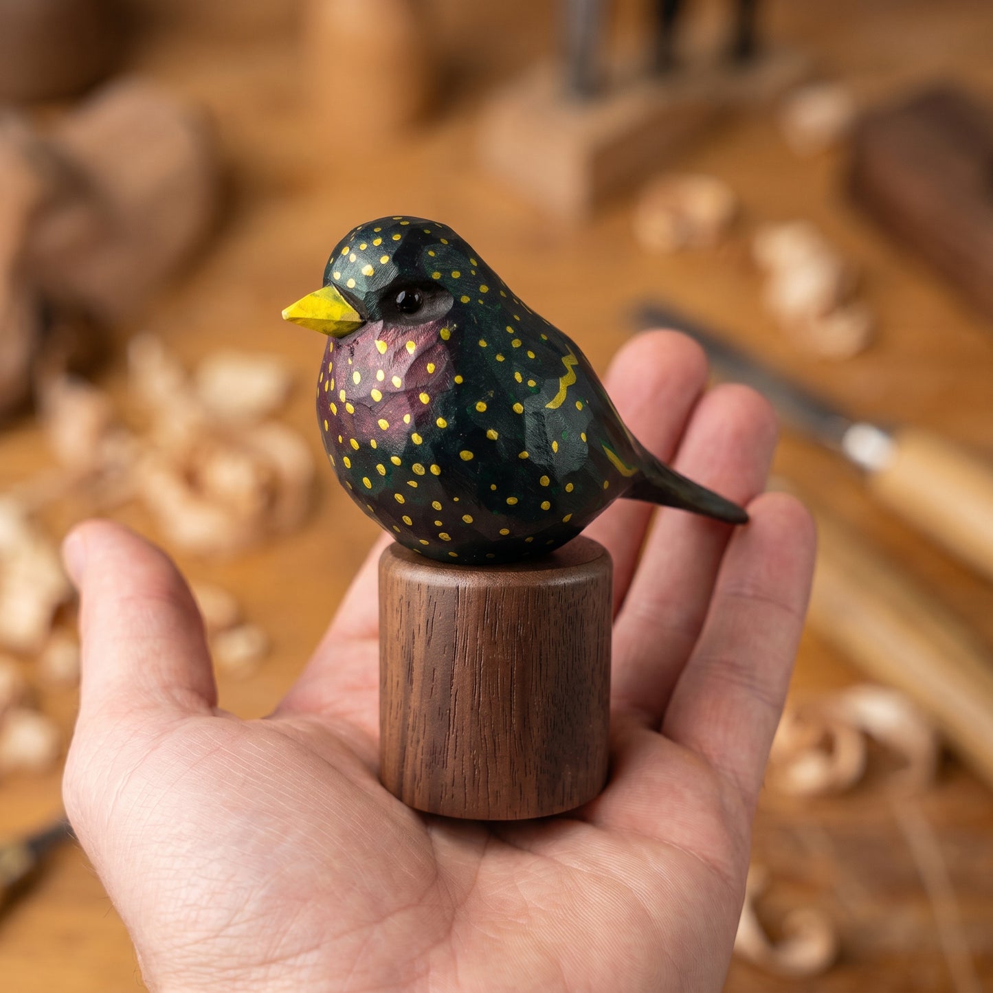 Hand holding a hand-carved wooden European Starling figurine on a round Black Walnut base in a woodcarving workshop