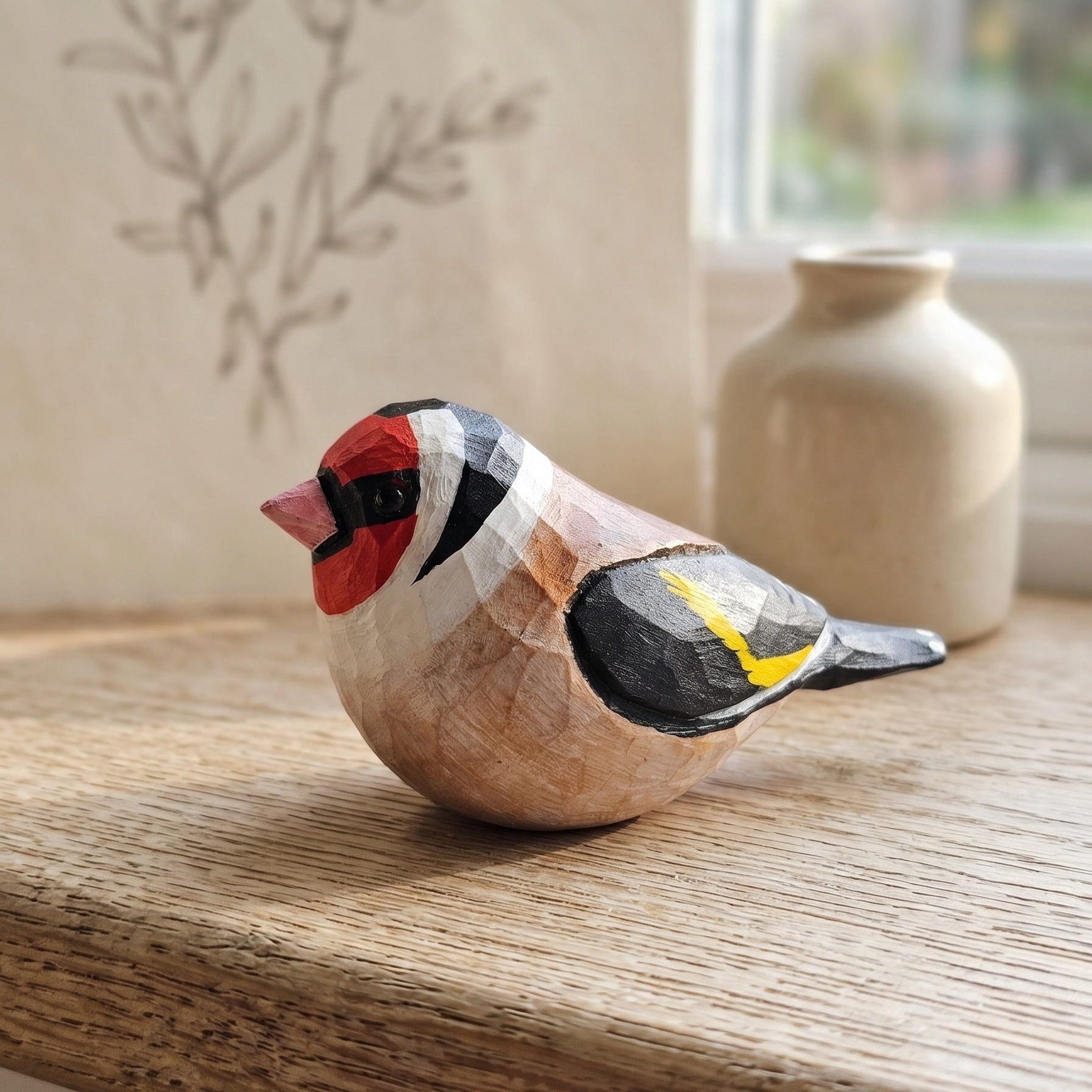 A hand-carved wooden European Goldfinch figurine resting on a rustic wooden windowsill, with a soft-focus beige ceramic vase and a window in the background.