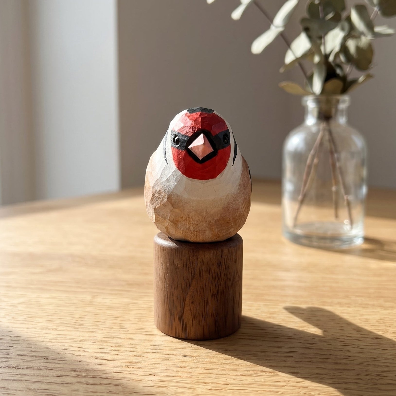 A handcrafted wooden European Goldfinch figurine with a red, black, and white painted face, mounted on a cylindrical wooden base and displayed on a light wood table next to a glass vase with eucalyptus.