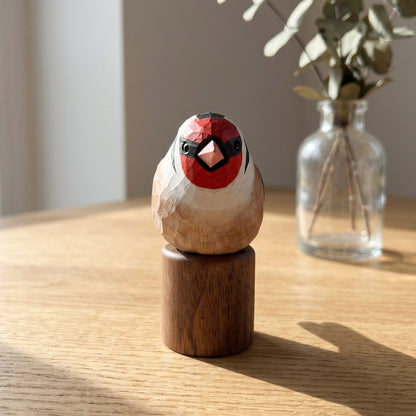 A handcrafted wooden European Goldfinch figurine with a red, black, and white painted face, mounted on a cylindrical wooden base and displayed on a light wood table next to a glass vase with eucalyptus.