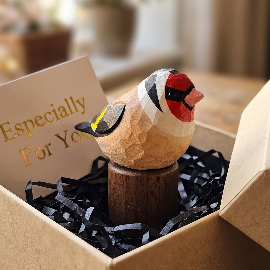 A small handcrafted wooden European Goldfinch figurine with red, black, and yellow painted details sits in a brown kraft gift box filled with black shredded paper and a card that reads 'Especially For You,' placed on a rustic wooden table in a softly blurred indoor setting.