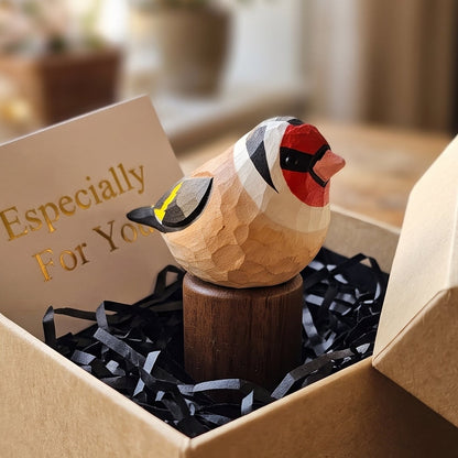 A small handcrafted wooden European Goldfinch figurine with red, black, and yellow painted details sits in a brown kraft gift box filled with black shredded paper and a card that reads 'Especially For You,' placed on a rustic wooden table in a softly blurred indoor setting.
