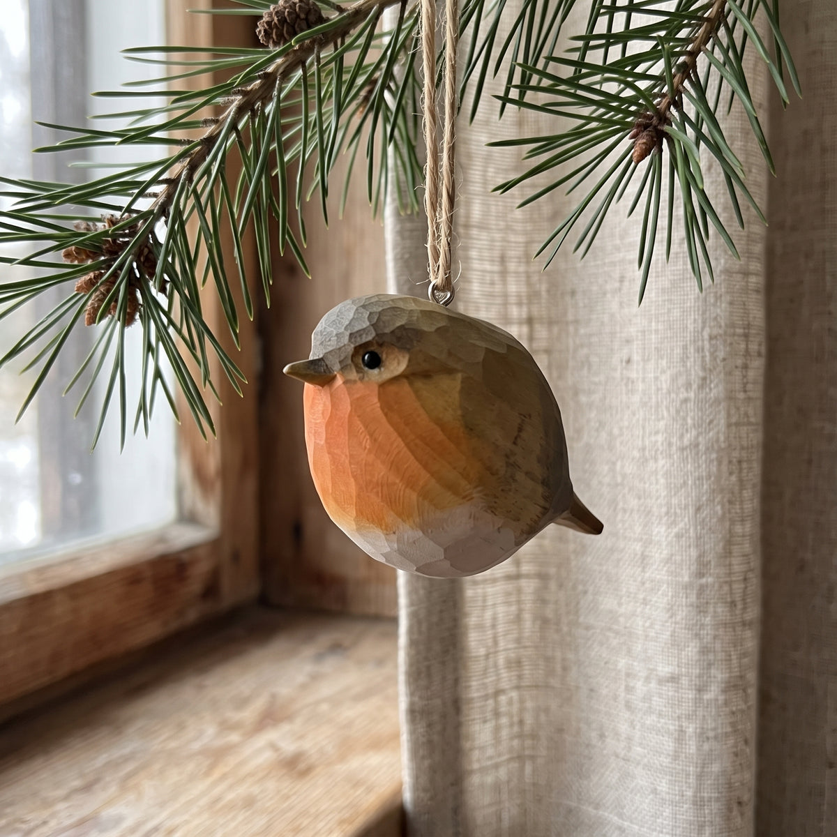 A faceted wooden robin bird ornament hanging from a pine branch in front of a window with beige curtains.