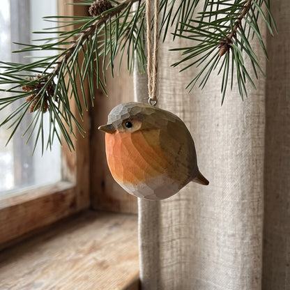 A faceted wooden robin bird ornament hanging from a pine branch in front of a window with beige curtains.