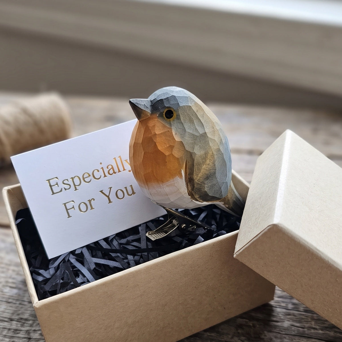 A hand-carved wooden European Robin clip-on ornament perched on the edge of an open kraft gift box filled with black shredded paper, featuring a white card reading "Especially For You" inside, set against a rustic wooden surface.
