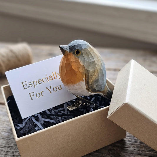 A hand-carved wooden European Robin clip-on ornament perched on the edge of an open kraft gift box filled with black shredded paper, featuring a white card reading "Especially For You" inside, set against a rustic wooden surface.