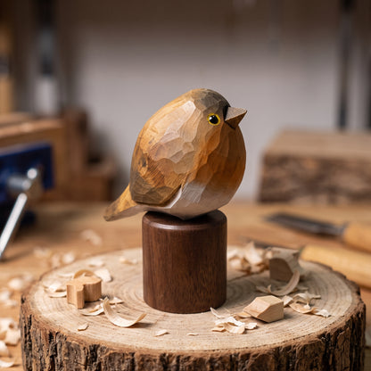 A carved wooden European robin figurine with a dark brown body and a lighter tan breast, mounted on a dark cylindrical pedestal. The bird is placed on a rustic wood slice with wood shavings scattered around, with a blurred workshop background showing tools and materials.