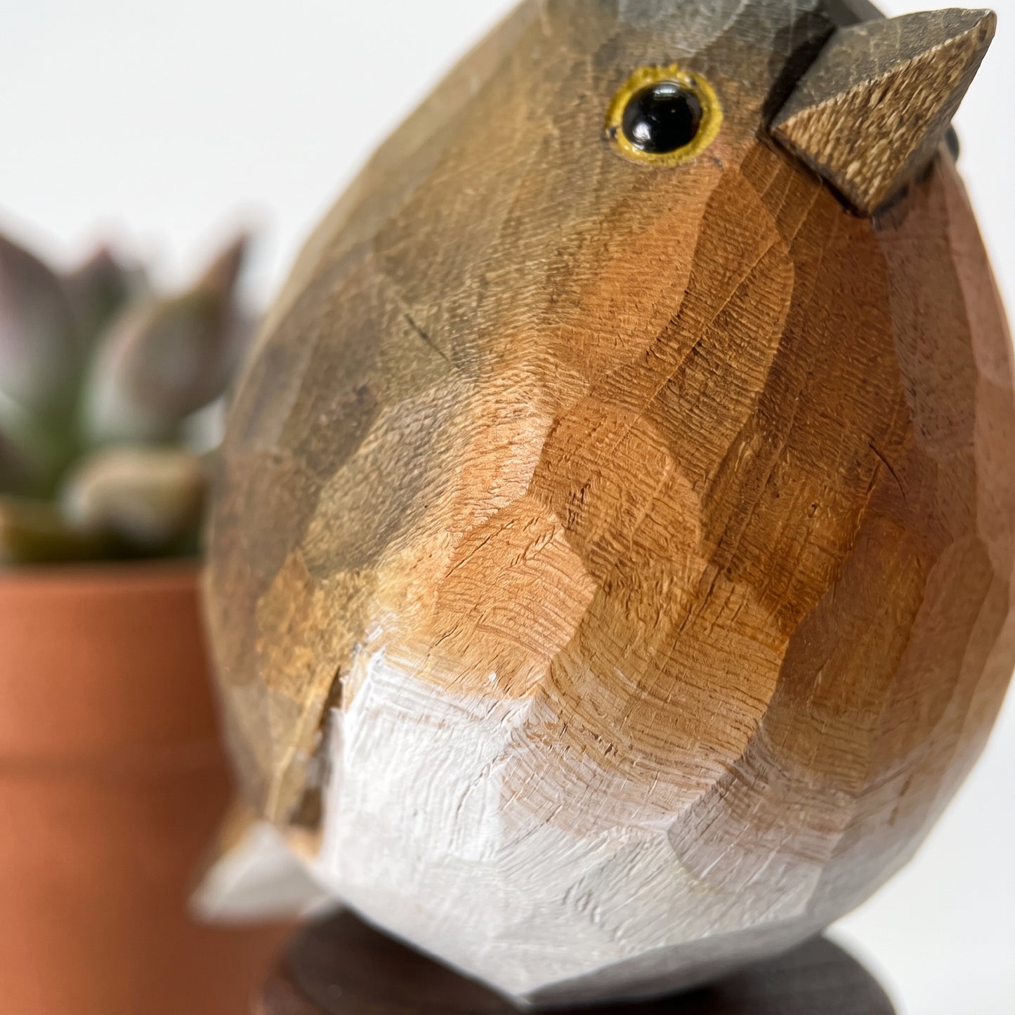 A close-up of a carved wooden robin bird figurine with a white belly, brown and tan back, and a small black eye with a yellow ring, shown against a softly blurred background with a terracotta pot.
