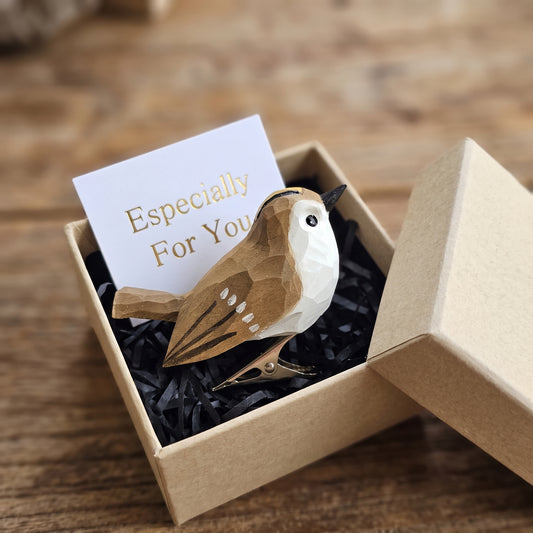 A hand-carved wooden Goldcrest clip-on ornament resting inside an open kraft gift box filled with black shredded paper, featuring a white cheek, olive-brown plumage, a silver metal clip underneath, and a white card reading "Especially For You" placed in the foreground on a rustic wooden tabletop.