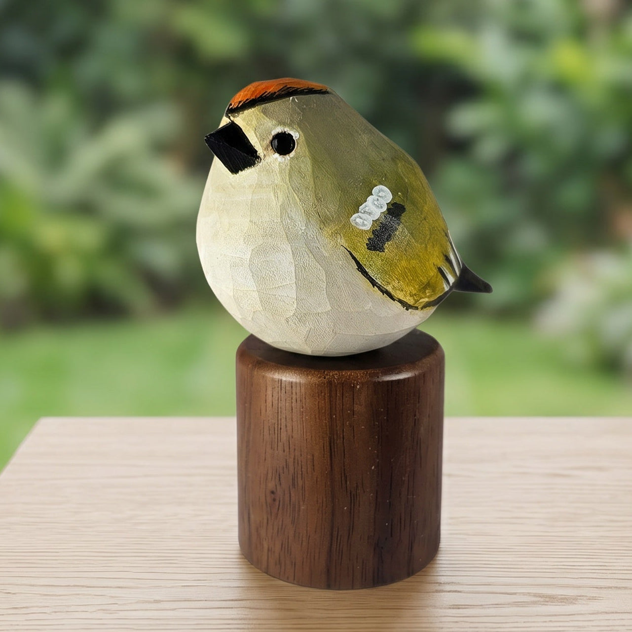 A carved wooden Golden-crowned kinglet figurine with a dark brown cylindrical base, displayed on a light wood table with a blurred green garden background.