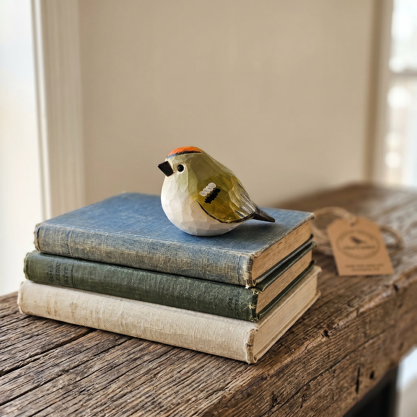 A small sculpted Golden-crowned kinglet figurine with olive green, white, black, and orange details sits on top of a stack of three vintage books on a rustic wooden table. A small paper tag is attached near the bird.